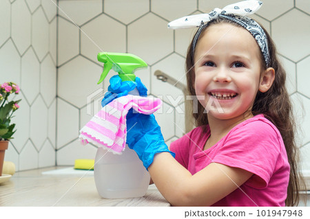Portrait of cute little preteen girl wearing big blue rubber gloves, holding sprayer, rag on white table for cleaning. 101947948