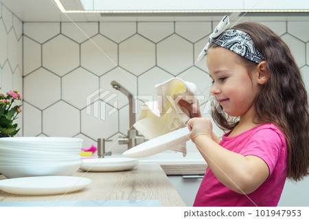 Side view of glad little girl with dark hair wearing pink T-shirt, washing dishes, drying clean white plates with cloth. Side view of glad little girl with dark hair wearing pink T-shirt, washing dishes, drying clean white plates with cloth. 101947953