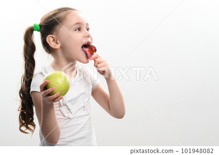 A little girl licks a lollipop and holds an apple in her hand. 101948082