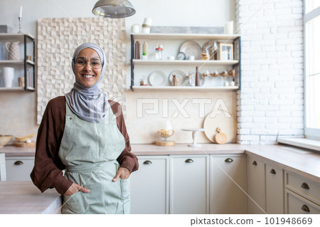 Portrait of Arab young housewife woman in hijab standing in kitchen near table in apron, holding hands in pockets, smiling and looking at camera. Portrait of Arab young housewife woman in hijab standing in kitchen near table in apron, holding hands in pockets, smiling and looking at camera. 101948669