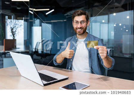 Bank credit card advertising. A young man sits in the office at the table and holds a golden money card, offers to buy, shows a super hand to the camera. Bank credit card advertising. A young man sits in the office at the table and holds a golden money card, offers to buy, shows a super hand to the camera. 101948683
