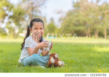 Caucasian mix girl drinking milk from glass sitting on grass at outdoor park, Whiskers from milk, Good nutrition of children concept. 101948786
