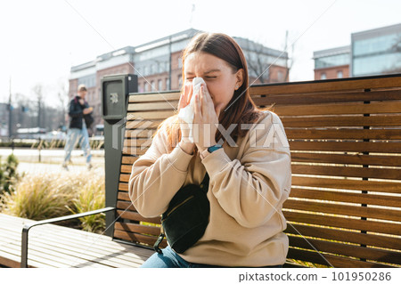 Unhappy caucasian millennial red-haired woman sneezing in napkin and siting in park bench, Young women suffering from allergy outdoors Unhappy caucasian millennial red-haired woman sneezing in napkin and siting in park bench, Young women suffering from allergy outdoors 101950286