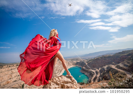 woman red dress lake mountains. Side view of a woman in a long red dress posing on a rock high above the lake. Against the background of the blue sky and the lake in the form of a heart woman red dress lake mountains. Side view of a woman in a long red dress posing on a rock high above the lake. Against the background of the blue sky and the lake in the form of a heart 101950295