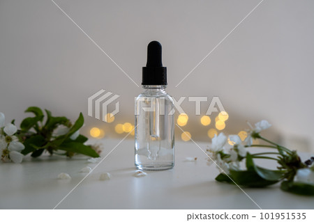Clear glass cosmetic dropper bottle and white cherry flowers with bokeh lights, care product mockup, faded colors Clear glass cosmetic dropper bottle and white cherry flowers with bokeh lights, care product mockup, faded colors 101951535