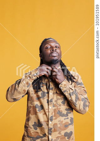 African american man buttoning up stylish shirt in front of camera in studio over yellow background. Cheerful young adult wearing fashionable clothes, preparing for elegant date 101951589