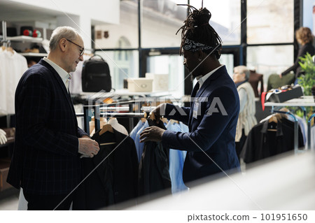 African american employee helping customer with suit in shopping centre, client looking to buy modern clothes. Elderly man buying fashionable merchandise in showroom. Fashion concept African american employee helping customer with suit in shopping centre, client looking to buy modern clothes. Elderly man buying fashionable merchandise in showroom. Fashion concept 101951650