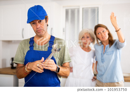 Inattentive plumber in uniform spreading his arms to the sides while being scolded by two women in the kitchen 101953750