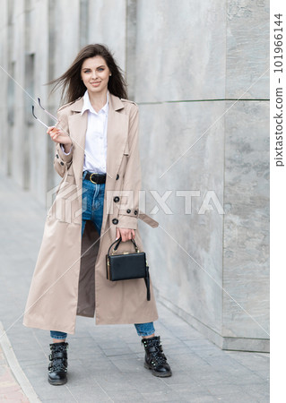 A young pretty girl in a raincoat and jeans walks around the city near a tall building, with a handbag in hand 101966144