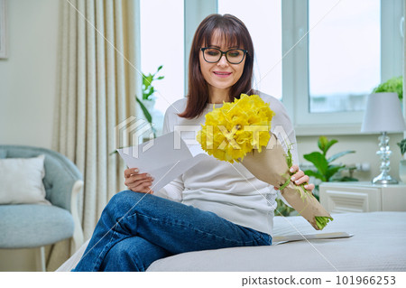 Happy middle aged female reading congratulations note, with bouquet of flowers, at home 101966253