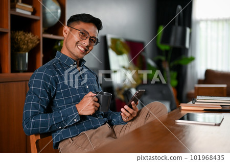 Successful Asian businessman sipping coffee and using his smartphone at his desk Successful Asian businessman sipping coffee and using his smartphone at his desk 101968435