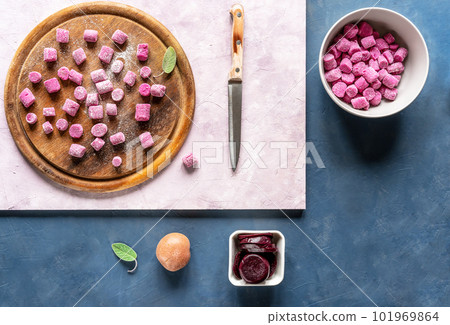 Beetroot and potato homemade gnocchi on round wooden cutting board and in a bowl, ingredients, sage leaf, knife, dark blue and pink backgrounds, top view 101969864