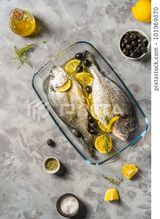 Two raw fresh sea bream or dorado are ready for cooking with lemon and spices in a glass casserole. Grey concrete background, napkin, top view. 101969870