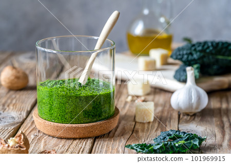 Homemade kale pesto served in a glass jar on wooden surface with grey background and basic ingredients - kale, walnuts, parmesan, garlic and olive oil Homemade kale pesto served in a glass jar on wooden surface with grey background and basic ingredients - kale, walnuts, parmesan, garlic and olive oil 101969915