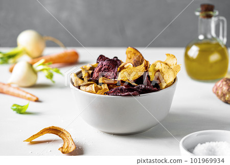 Healthy vegetable chips in a bowl over light surface with grey concrete background. Marine salt, raw vegetables and oil aside Healthy vegetable chips in a bowl over light surface with grey concrete background. Marine salt, raw vegetables and oil aside 101969934
