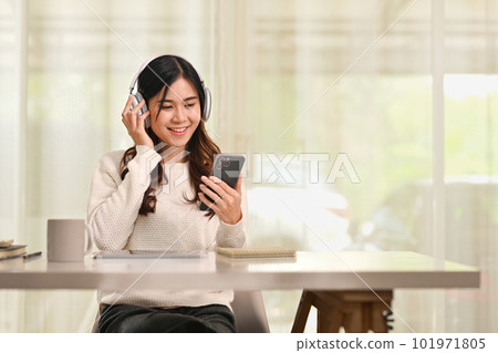 Cheerful asian woman listening to music in wireless headphones and using smartphone at desk in living room 101971805