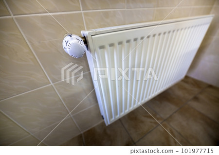 Close-up of white heating radiator with thermostat valve ion tiled walls and floor background. Comfortable warm home interior, climate control, money saving concept. 101977715