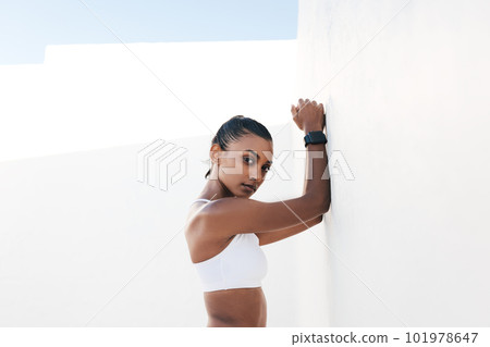 Portrait of a fit female leaning white wall and looking straight at a camera. Fitness influencer taking a short break during a workout. 101978647