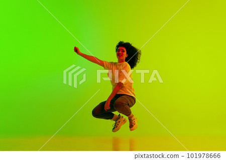 Young girl, hip-hop dancer in sport style clothes jumping against gradient green yellow background in neon light Young girl, hip-hop dancer in sport style clothes jumping against gradient green yellow background in neon light 101978666