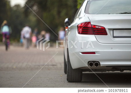 Back view part of white car parked on city pedestrian zone pavement on background of blurred silhouettes of biker and people walking along green sunny summer alley. Modern city lifestyle concept. 101978911