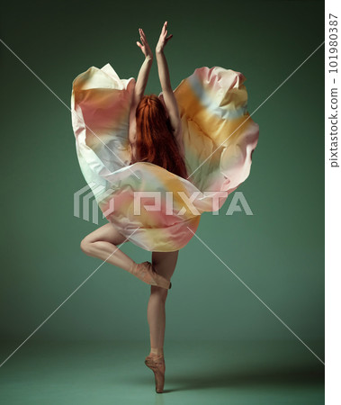 Young classical dancer wearing colorful flying dress dancing on fingertips over dark green studio background. Tender movement 101980387