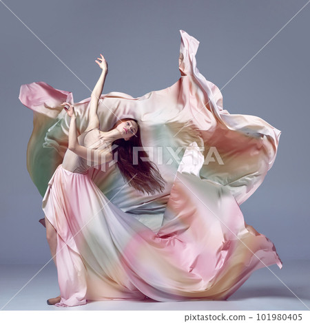 Inspired young, beautiful ballerina wearing rainbow dress dancing over grey studio background. Ballet with silk dress 101980405