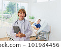 the doctor is examining the patient in the hospital. Caucasian female doctor in a gown Hand holding tablet looking at camera with patient in background in hospital. 101984456