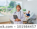the doctor is examining the patient in the hospital. Caucasian female doctor in a gown Hand holding tablet looking at camera with patient in background in hospital. 101984457