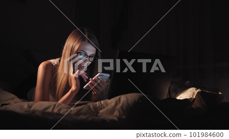 Side view of a happy teen browsing and watching media in a smart phone lying on a bed in the night at home. Side view of a happy teen browsing and watching media in a smart phone lying on a bed in the night at home. 101984600