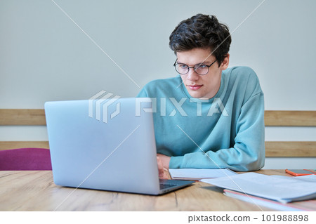 Young guy college student sitting at desk in classroom, using laptop, writing in notebook Young guy college student sitting at desk in classroom, using laptop, writing in notebook 101988898