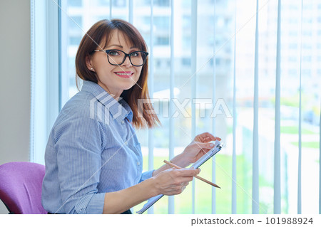 Middle aged woman with clipboard sitting in office near window. Middle aged woman with clipboard sitting in office near window. 101988924