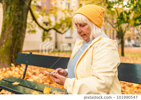 Senior 50s woman walking outdoors in autumn spring park using mobile phone. 101990547