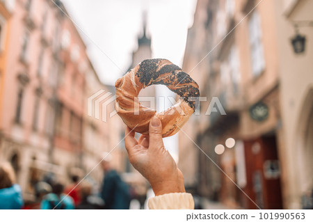 Happy young blonde woman tourist in stylish clothes holding pretzel obwarzanek on the market square in Krakow in Poland 101990563