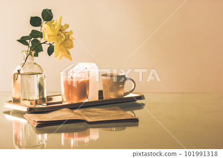 Still life in beige tones with a stylish tray, a cup of tea or coffee, a blooming yellow rosebud in a glass vase, an old open book lying on a table. Modern home decor for interior. Copy space for text 101991318