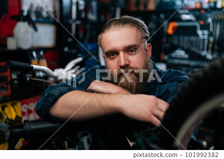 Closeup face of bearded cycling repairman standing leaning on bicycle in repair bike workshop with dark interior, serious looking at camera. Concept of professional repair and maintenance of bicycle. Closeup face of bearded cycling repairman standing leaning on bicycle in repair bike workshop with dark interior, serious looking at camera. Concept of professional repair and maintenance of bicycle. 101992382