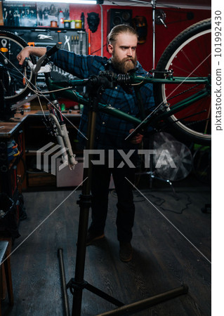 Vertical portrait of repairman watching rear shifter of mountain bike, changing speeds using handlebar shift lever working in bicycle repair shop with dark interior. Concept of maintenance of bicycle. 101992430