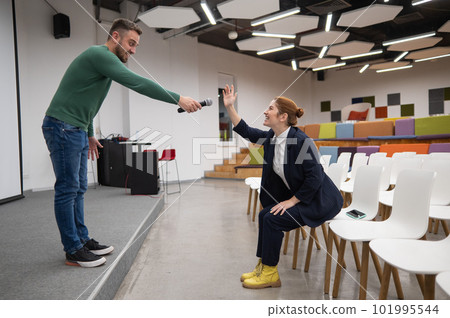 A red-haired Caucasian business woman sits in the front row in an empty conference room. Bearded man asks a question in the hall.  101995544