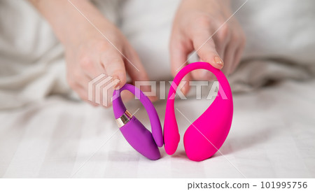 Woman holding pink and purple vibrators while lying in bed.  101995756