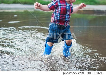 Happy toddler boy running through puddles in rubber boots. Child splashes after rain outside.. 101999693