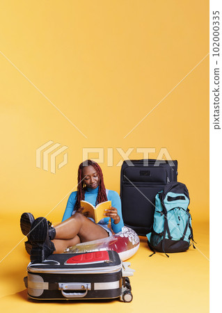 African american tourist reading book, sitting on the floor with inflatable and baggage in studio. Young woman enjoying lecture for leisure activity, travelling abroad on citybreak. African american tourist reading book, sitting on the floor with inflatable and baggage in studio. Young woman enjoying lecture for leisure activity, travelling abroad on citybreak. 102000335