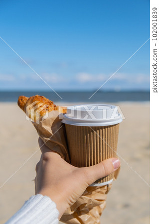 Tasty fresh croissants with coffee in paper cup in female hands on beach sea background. Breakfast outdoors wellbeing concept. FPV. Take away food Tasty fresh croissants with coffee in paper cup in female hands on beach sea background. Breakfast outdoors wellbeing concept. FPV. Take away food 102001839