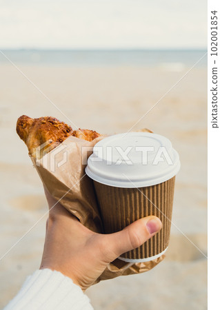 Tasty fresh croissants with coffee in paper cup in female hands on beach sea background. Breakfast outdoors wellbeing concept. FPV. Take away food Tasty fresh croissants with coffee in paper cup in female hands on beach sea background. Breakfast outdoors wellbeing concept. FPV. Take away food 102001854