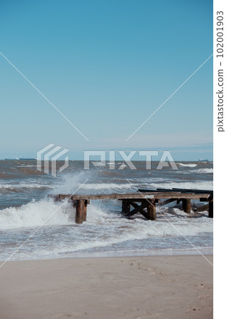Storm sea waves empty wooden pier in dramatic blue sky background. Seascape with running splashing sea waves. Sandy beach vacation getaway in Gdansk Poland.  102001903