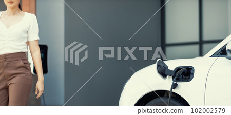 Focus EV car at home charging station with blur progressive woman in the back. 102002579