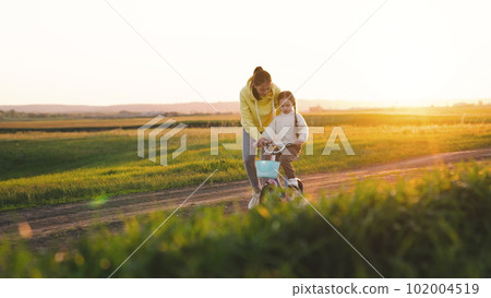 Happy mother teaches her little daughter how to ride a two-wheeled bicycle. family childhood dream. sunlight child bike riding. mother child kid girl outdoors. child rides bike for first time sunset 102004519