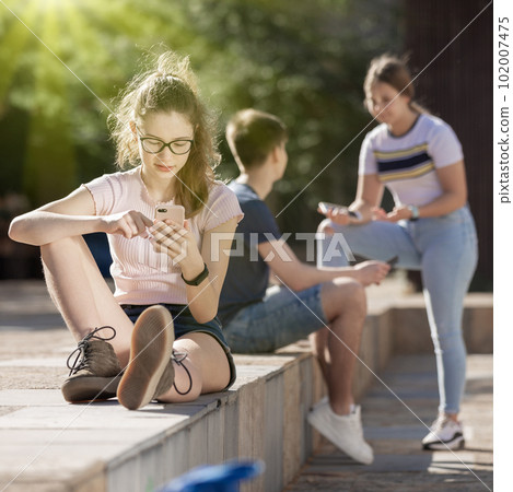 Teen girl playing on smartphone sitting near to friends Teen girl playing on smartphone sitting near to friends 102007475