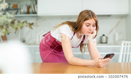 Concentrated European teenage girl in red polka dot apron using mobile phone and surfing Internet in cozy cuisine Concentrated European teenage girl in red polka dot apron using mobile phone and surfing Internet in cozy cuisine 102007476