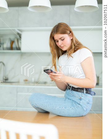 Concentrated European teenage girl in casual clothes searching info or recipe on smartphone while sitting on kitchen table 102007511