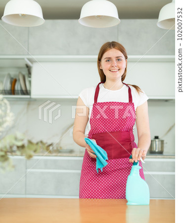 in kitchen,positive woman hostess in dotted red apron stands and holds rag and cleaning spray 102007532