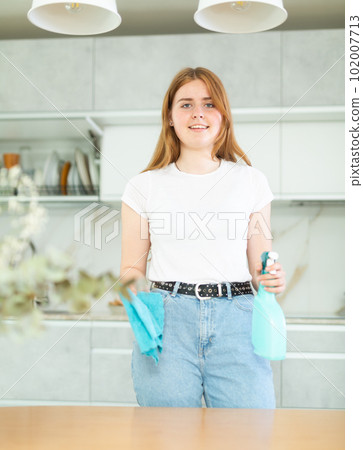 Joyful smiling teen girl holding spray and microfiber rag before cleaning cozy kitchen 102007713